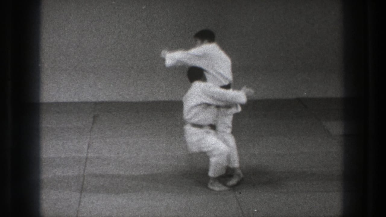 One adult man in a white gi lifts another on a mat during a throw in the Tokyo martial-arts footage.