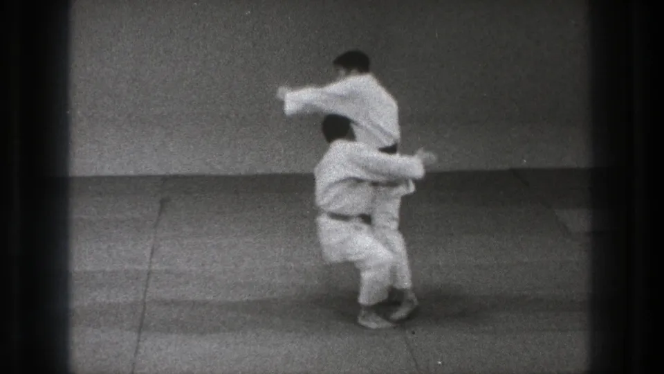 One adult man in a white gi lifts another on a mat during a throw in the Tokyo martial-arts footage.