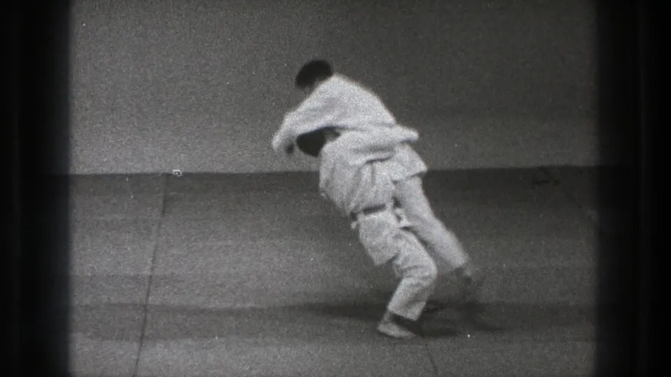 In supported 1971 Tokyo footage, two adult practitioners in white uniforms engage at the start of a martial arts throw on an indoor mat.