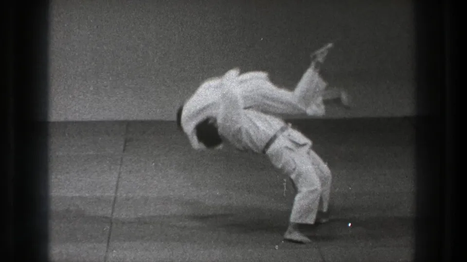 A white-uniformed practitioner lifts an opponent over his back during a slow-motion martial arts throw in the Tokyo 1971 clip.
