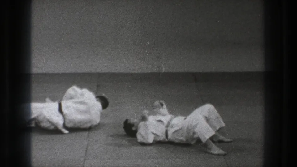 In 1971 Tokyo footage, two adult men in white gi and dark belts are down on a dojo mat during a martial arts drill.