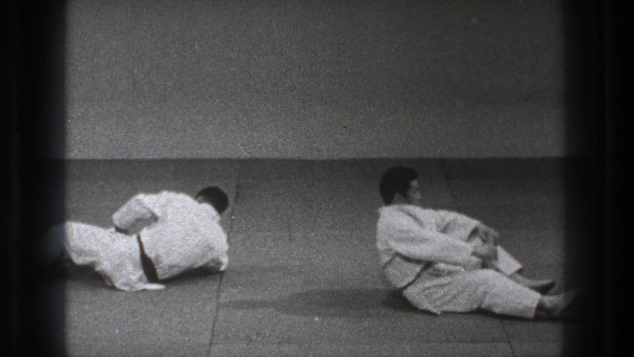 Two adult practitioners in white gi continue a floor movement on a plain dojo mat in the supplied 1971 Tokyo clip.