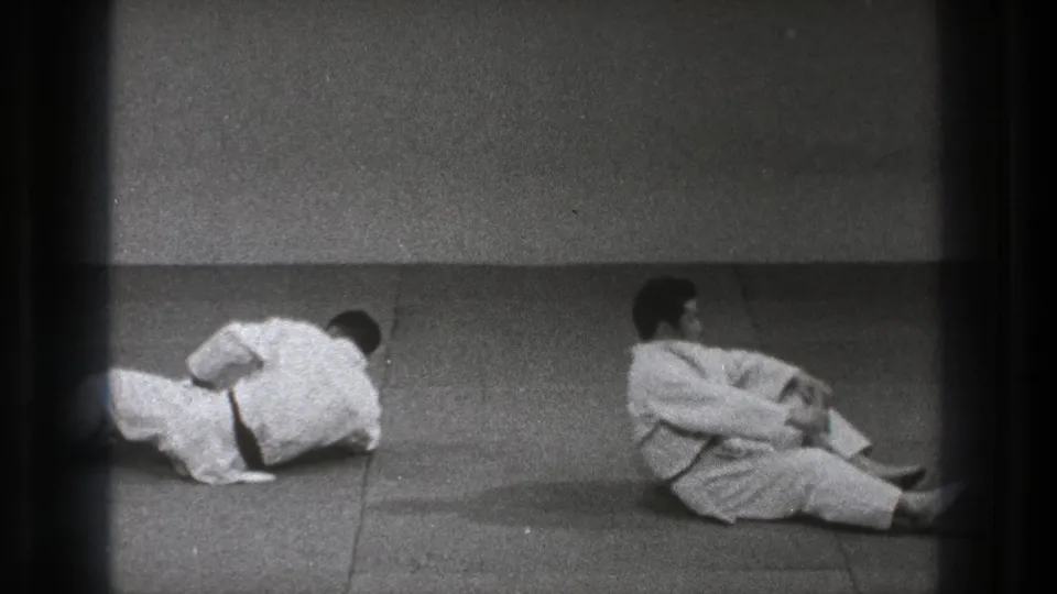 Two adult practitioners in white gi continue a floor movement on a plain dojo mat in the supplied 1971 Tokyo clip.