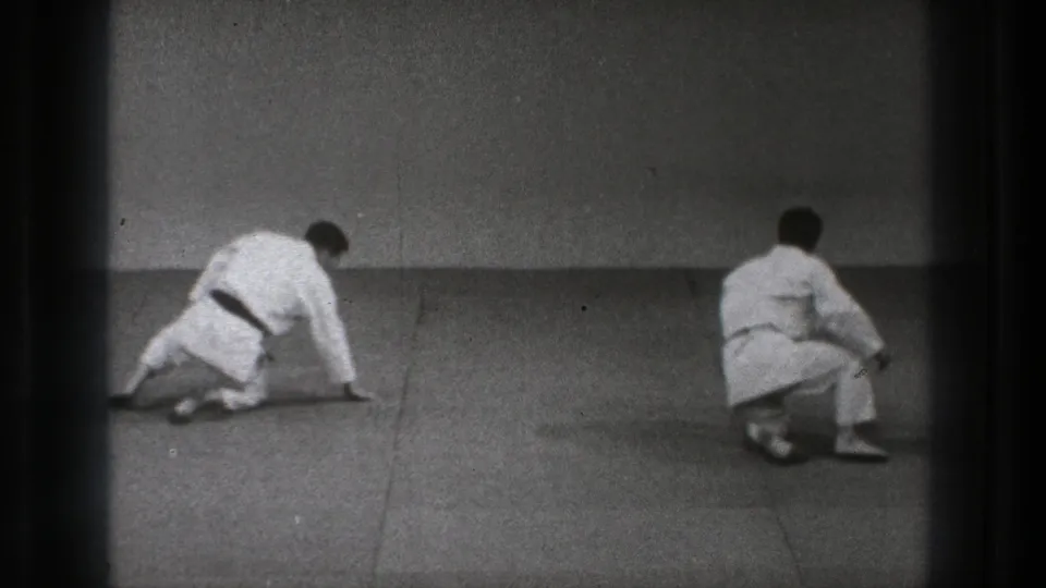 The two men move toward upright positions on the mat, with one kneeling and the other seated during the martial arts practice.