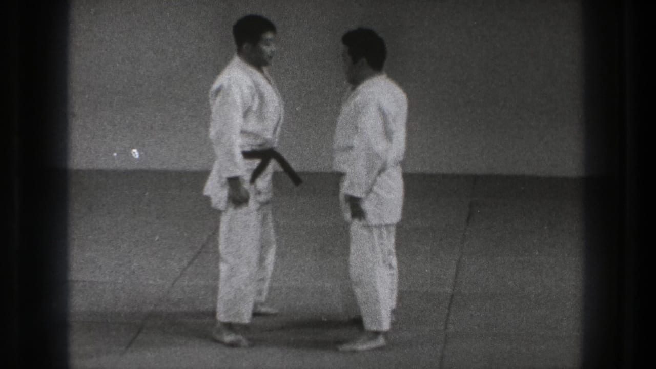 The two adult martial arts practitioners stand chest-to-chest on the indoor mat during the setup for Sumi Gaeshi.