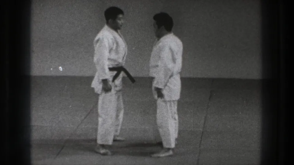 The two adult martial arts practitioners stand chest-to-chest on the indoor mat during the setup for Sumi Gaeshi.