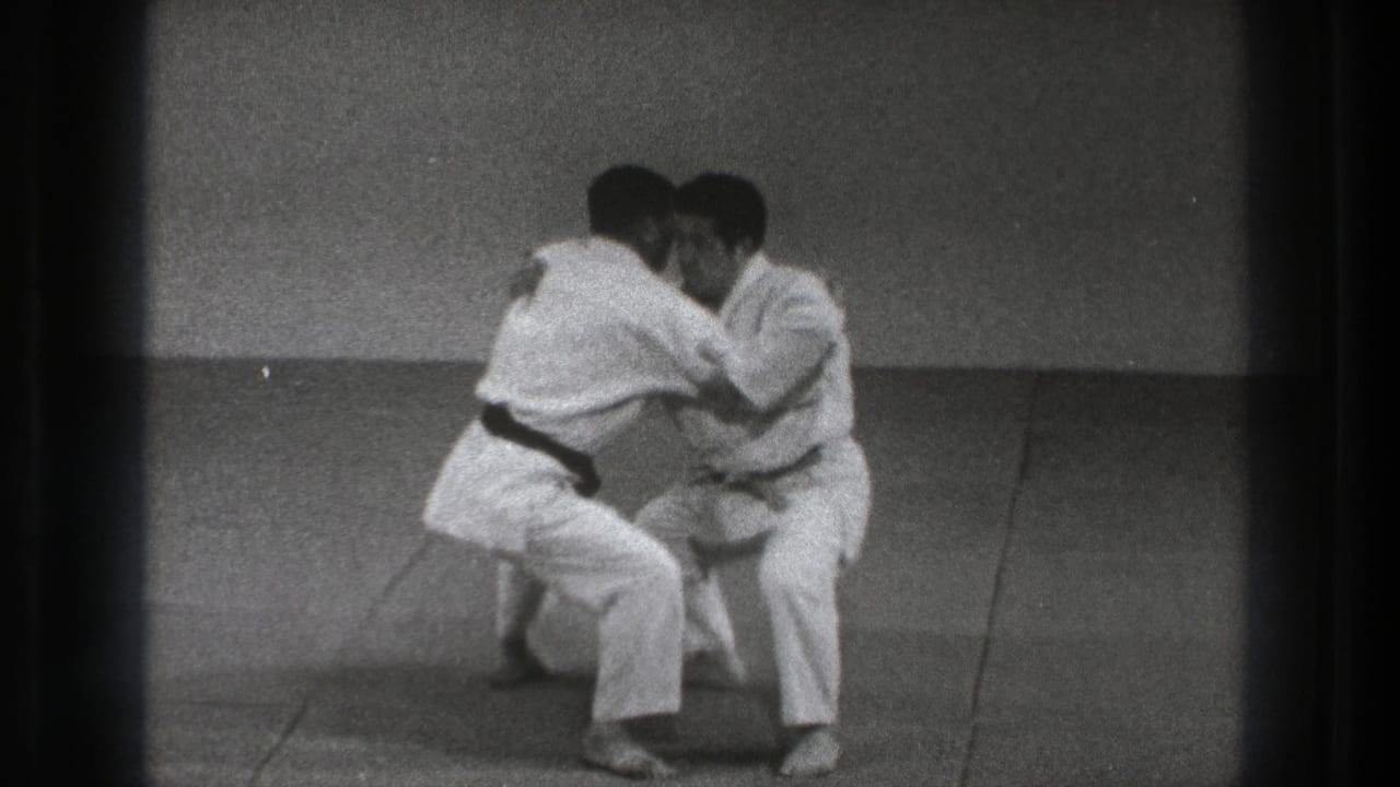 In 1971 Tokyo footage, two adult karate practitioners in white gi crouch on an indoor mat during a defensive grab drill.