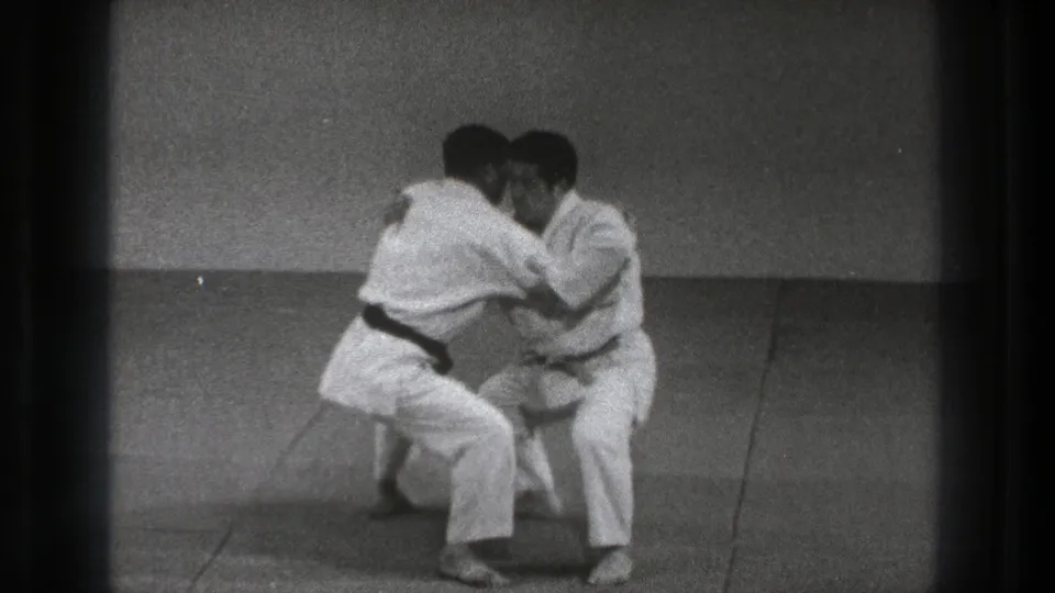 In 1971 Tokyo footage, two adult karate practitioners in white gi crouch on an indoor mat during a defensive grab drill.