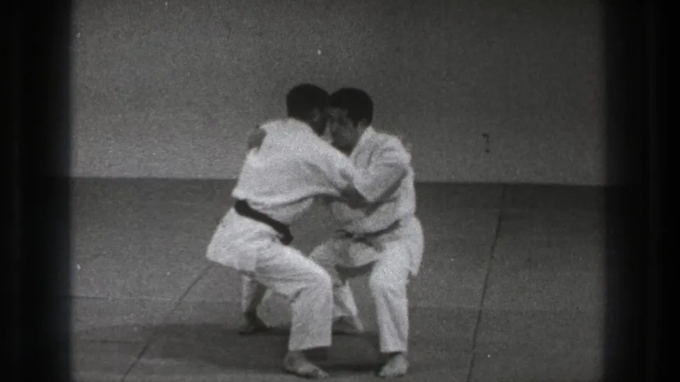 Two adult men in white gi practice a close-contact karate defense on a plain indoor mat in the Tokyo 1971 clip.