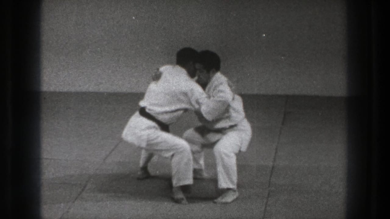 The 1971 Tokyo karate practice continues with both men low to the mat, hands engaged in a defensive grab drill.