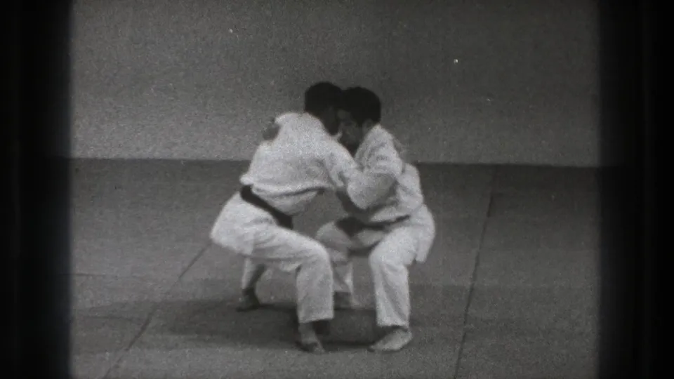 The 1971 Tokyo karate practice continues with both men low to the mat, hands engaged in a defensive grab drill.