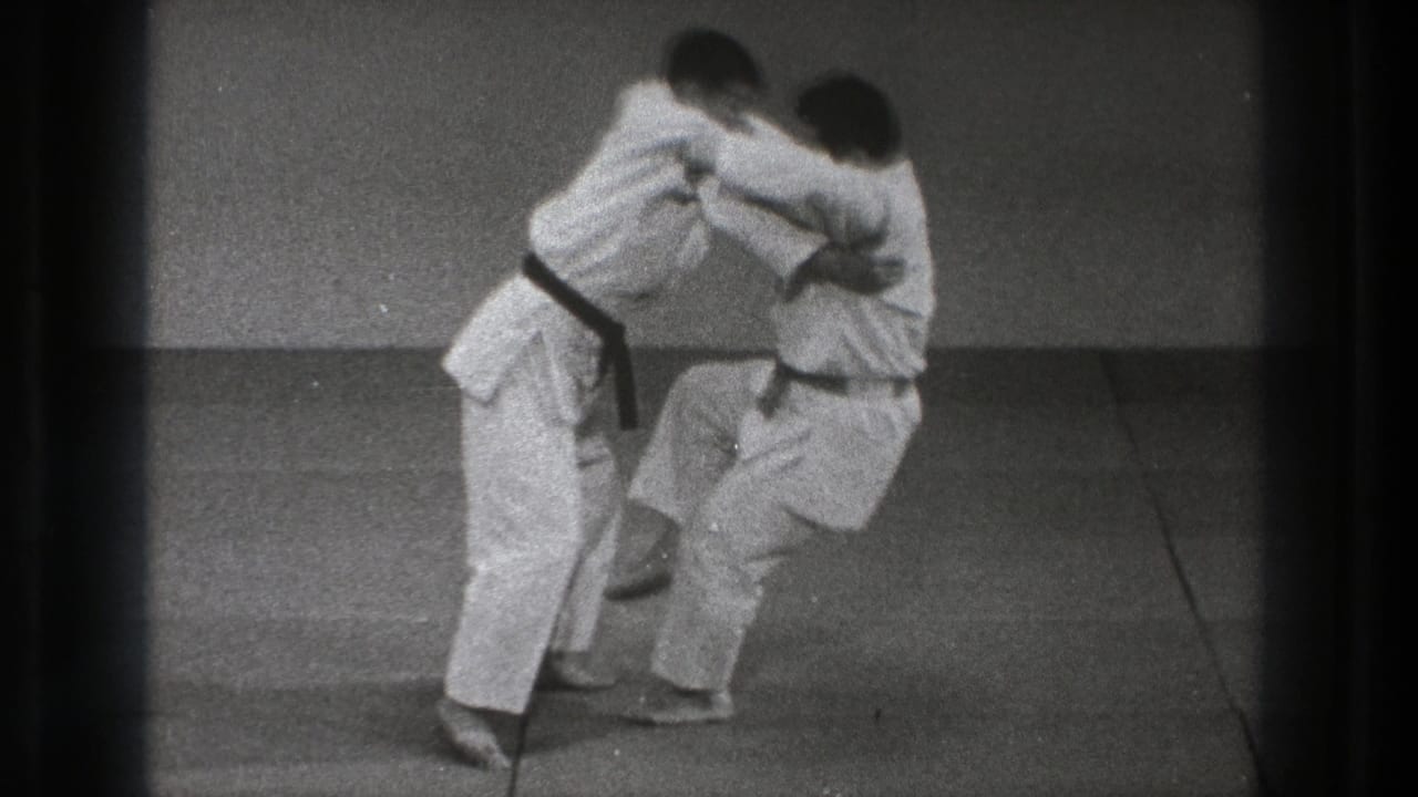 Two martial artists in white uniforms grapple on a grey mat.