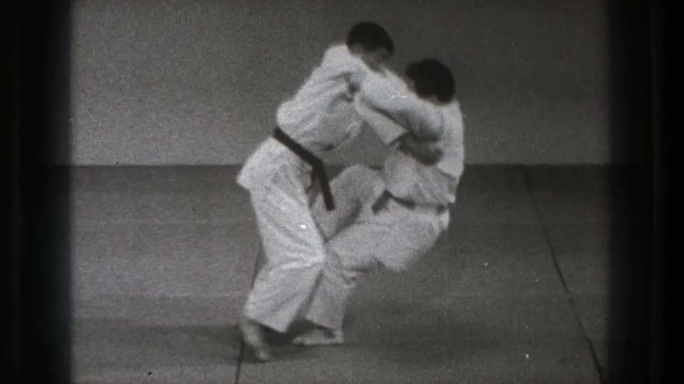 A black-belt practitioner lifts his partner during a martial arts throw in the 1971 Tokyo dojo footage.