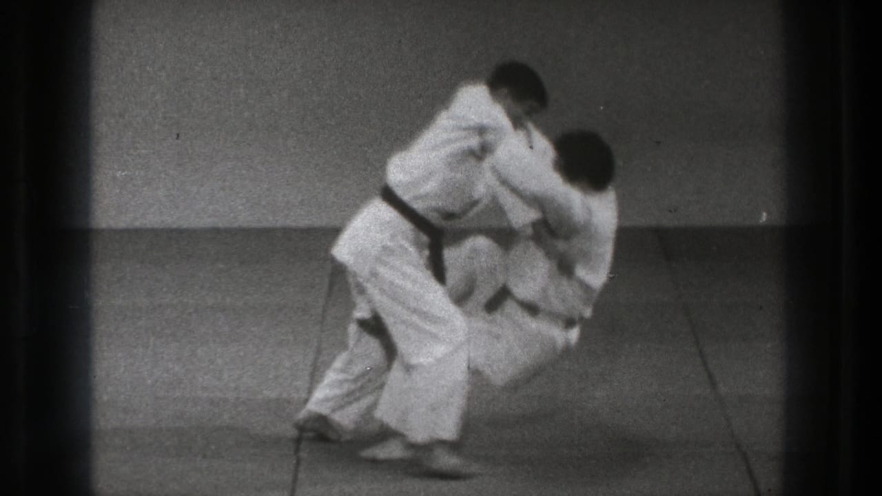The throw continues as one practitioner bends low while the other is pulled forward on the grey mat.