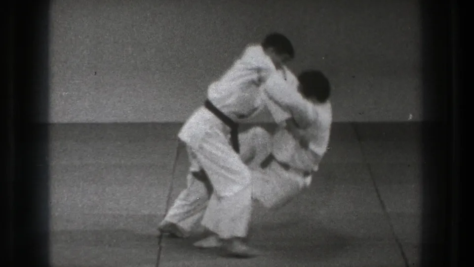 The throw continues as one practitioner bends low while the other is pulled forward on the grey mat.