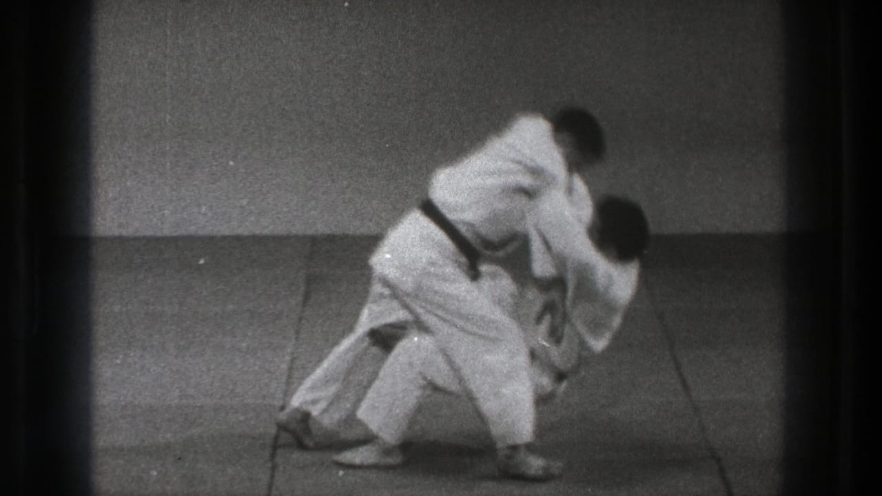 In Tokyo 1971 footage, two judo practitioners in white gi begin a throw on a tatami mat inside a plain dojo.