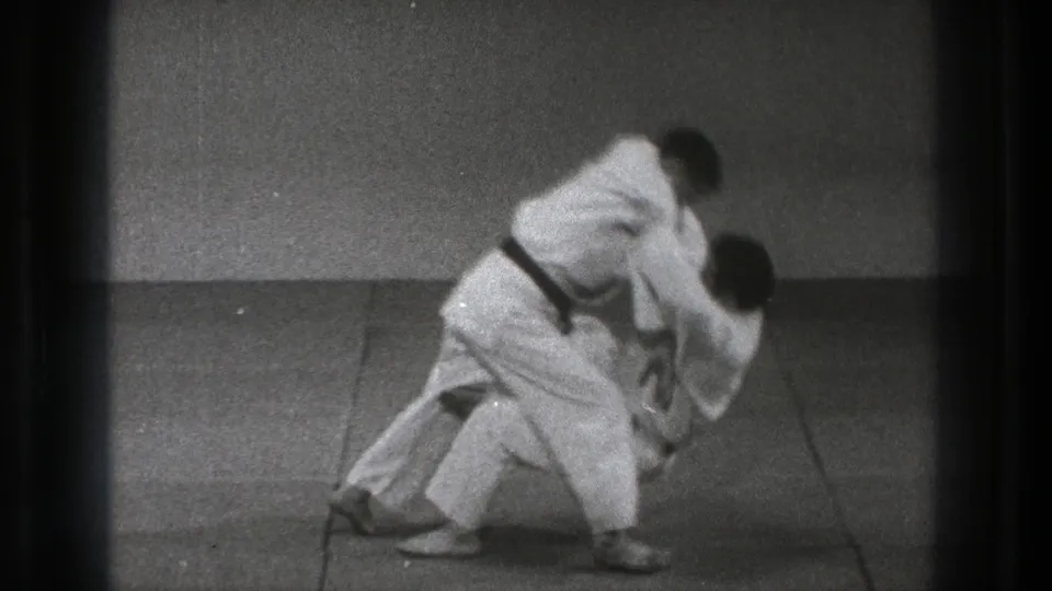 In Tokyo 1971 footage, two judo practitioners in white gi begin a throw on a tatami mat inside a plain dojo.