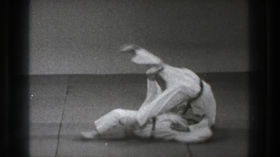 After the throw, one practitioner lies on the tatami while the other remains low and close in the dojo practice space.