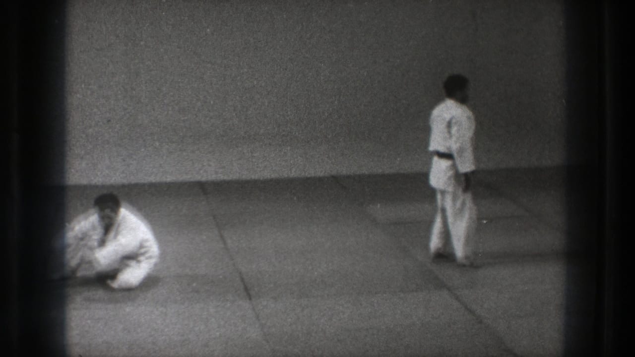 Two men in karate uniforms on a mat, with one seated and one standing nearby.