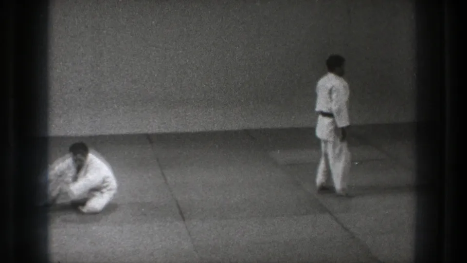 1971 Tokyo 8mm frame showing two karate practitioners in white gi on a plain dojo mat, one seated and one standing.