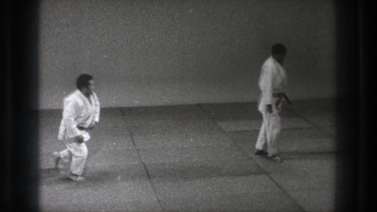1971 Tokyo dojo footage with one man in a white gi getting up from the mat as a black-belt partner stands opposite.