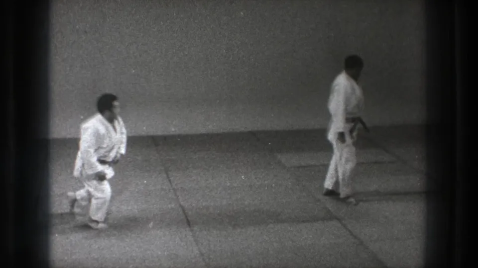 1971 Tokyo dojo footage with one man in a white gi getting up from the mat as a black-belt partner stands opposite.