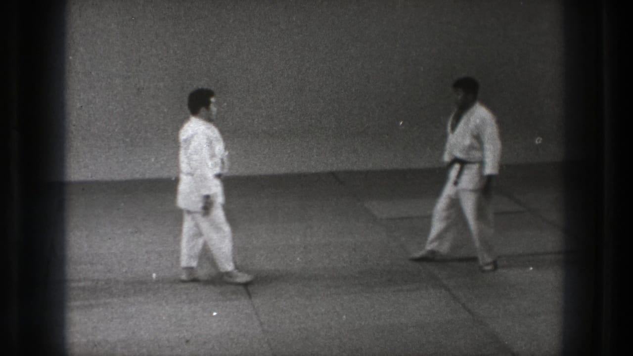 1971 Tokyo 8mm frame of two men in karate gi approaching or facing each other on a plain indoor training mat.