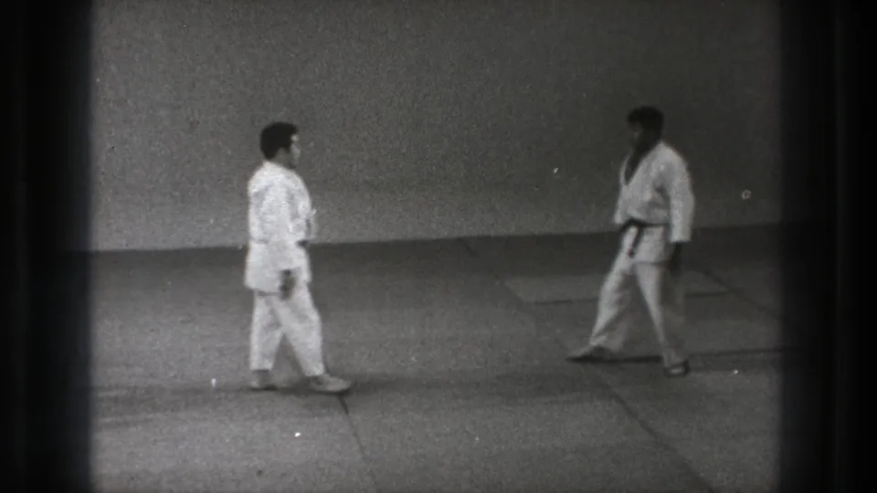 1971 Tokyo 8mm frame of two men in karate gi approaching or facing each other on a plain indoor training mat.