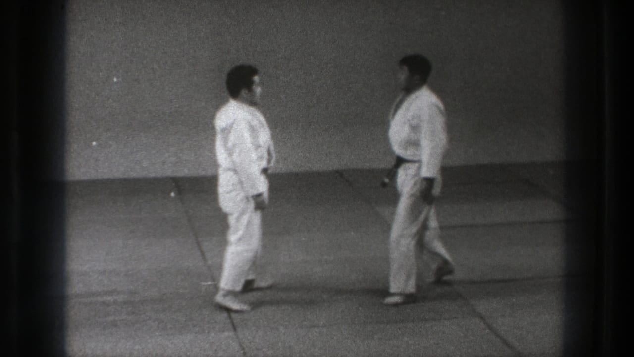 1971 Tokyo home-movie frame showing two karate practitioners in white gi standing face to face in a sparse dojo.