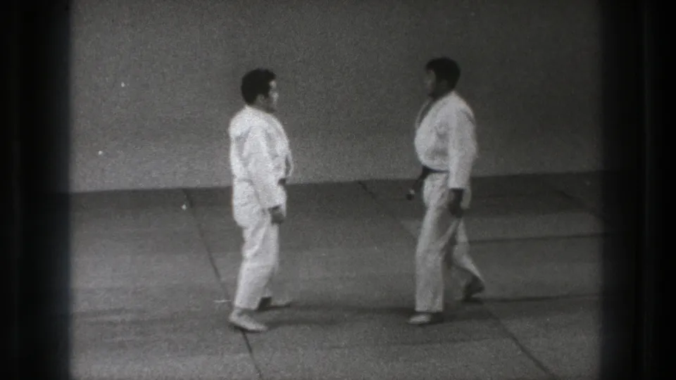 1971 Tokyo home-movie frame showing two karate practitioners in white gi standing face to face in a sparse dojo.