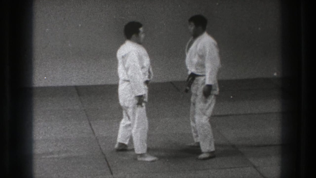 Two men in white martial arts uniforms stand facing each other on a grey mat.
