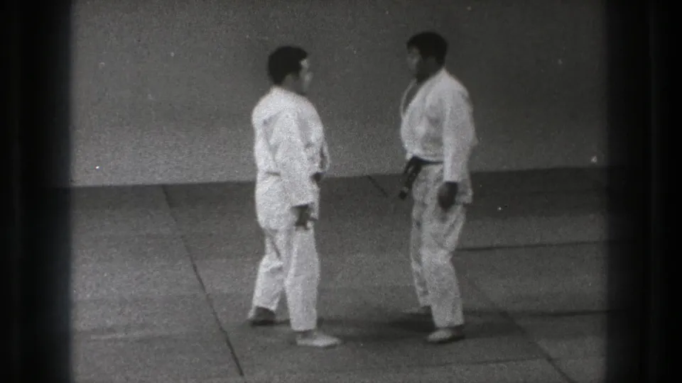 In 1971 Tokyo footage, two adult men in white martial arts uniforms face each other on a simple indoor mat before beginning a drill.