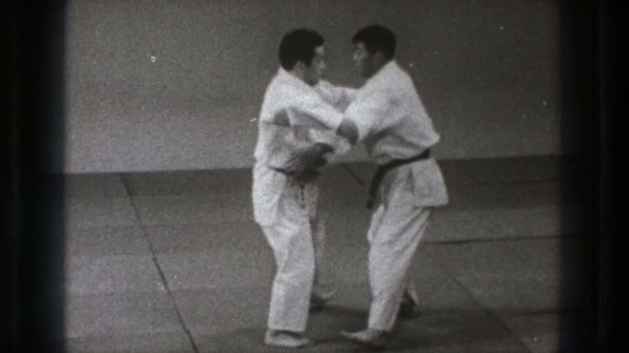 Two adult men in white martial arts uniforms make close arm contact during a standing technique in the Tokyo dojo footage.