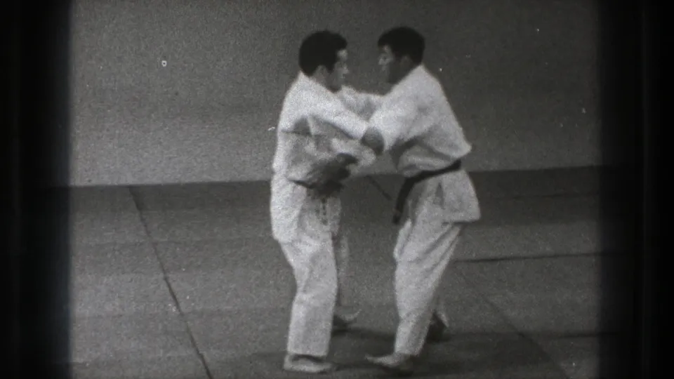 Two adult men in white martial arts uniforms make close arm contact during a standing technique in the Tokyo dojo footage.
