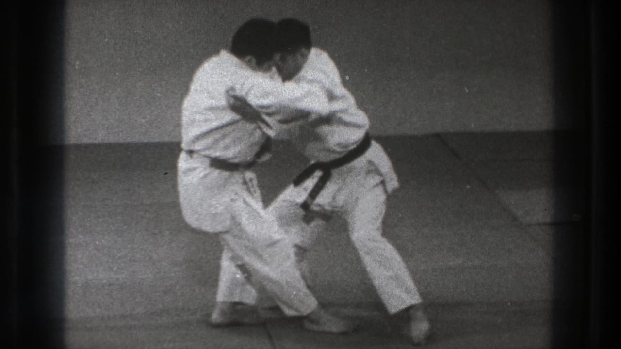 Two adult practitioners in white uniforms lean into a controlled close hold on the dojo mat near the end of the short 1971 Tokyo clip.