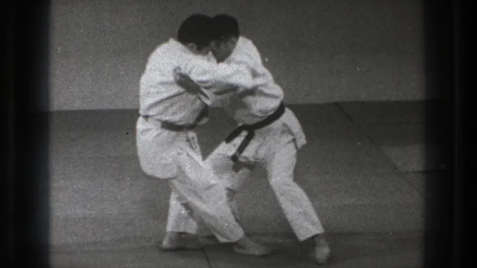 Two adult practitioners in white uniforms lean into a controlled close hold on the dojo mat near the end of the short 1971 Tokyo clip.