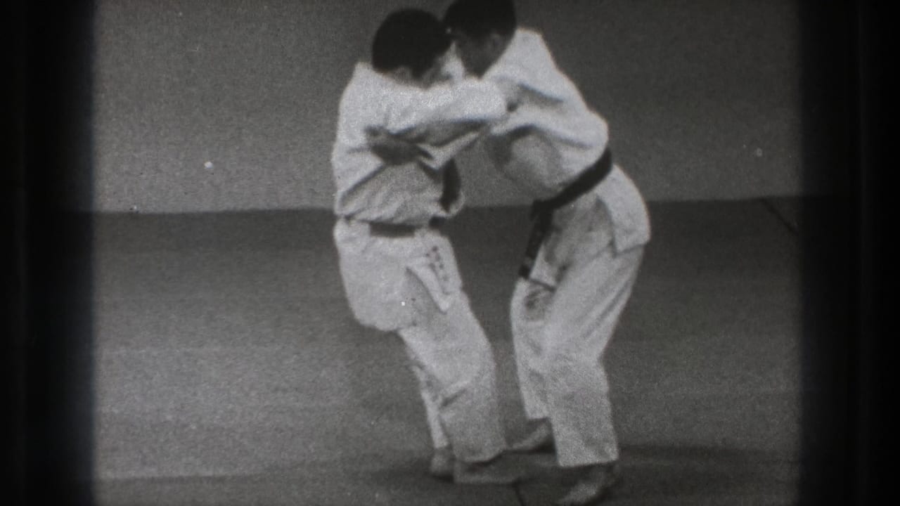 Two martial artists in white gi grip each other on an indoor mat.