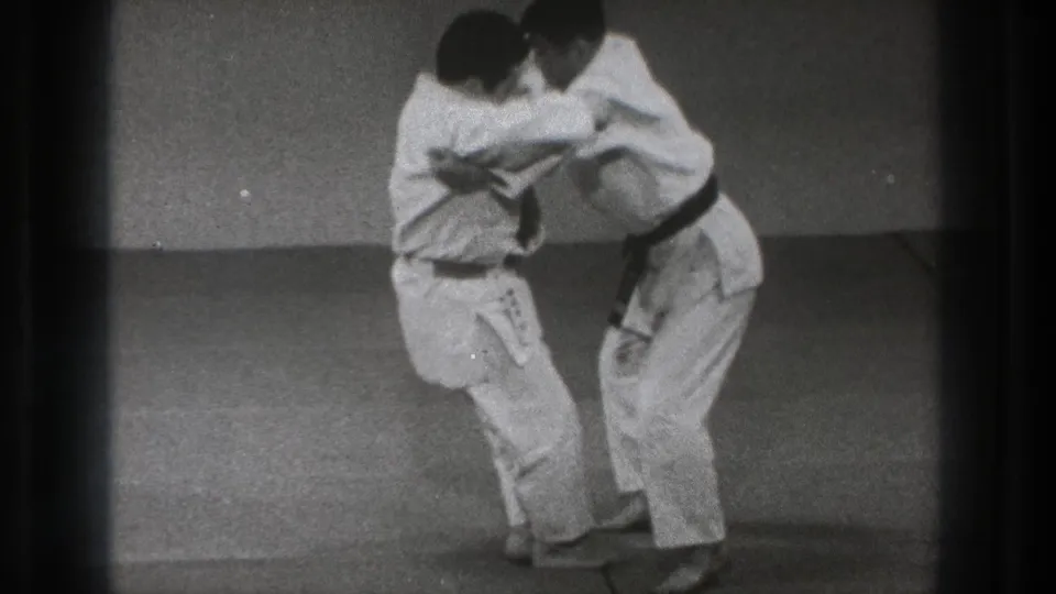 Two martial artists in white gi practice a standing grip in 1971 Tokyo indoor training footage.