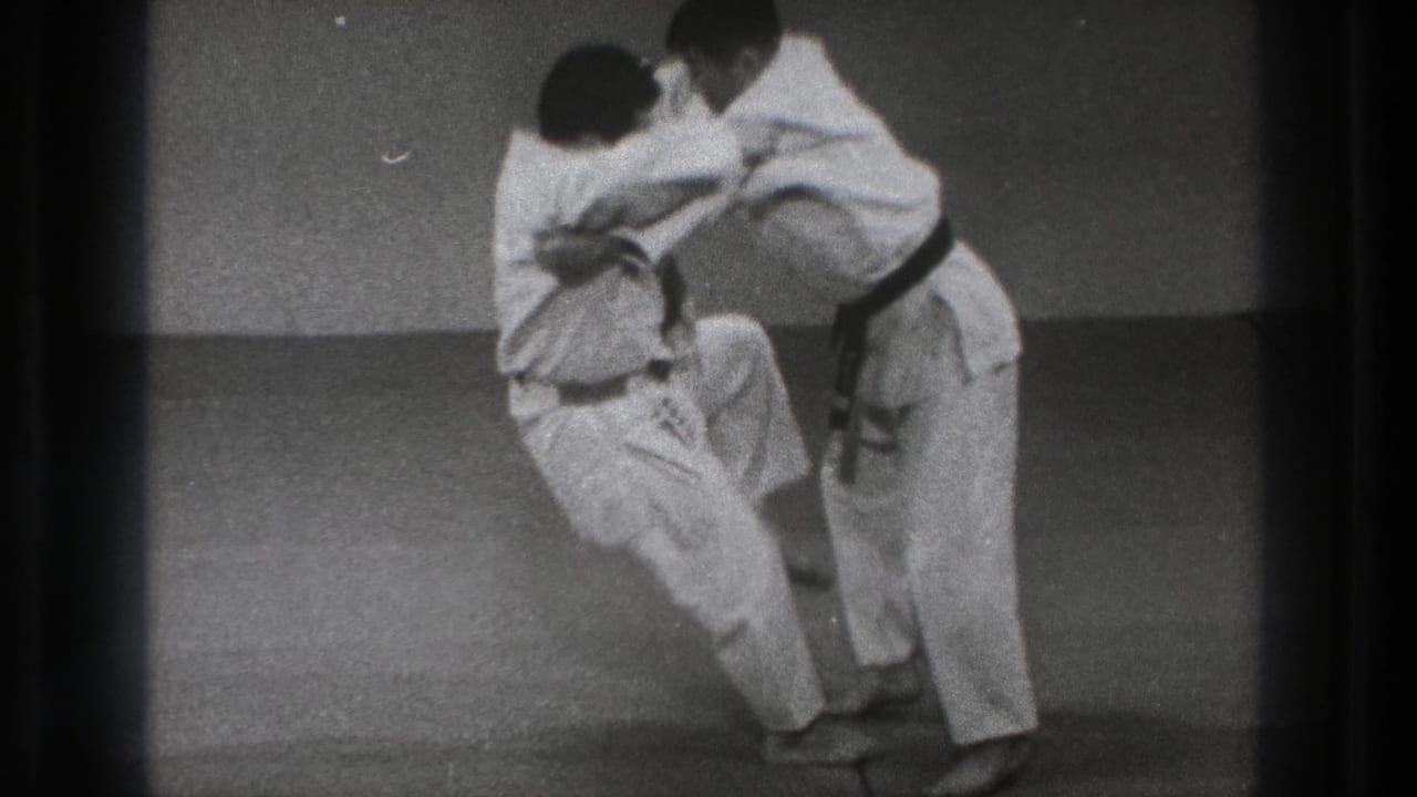 During the Tokyo practice sequence, one martial artist shifts balance and raises a knee while maintaining the grip.