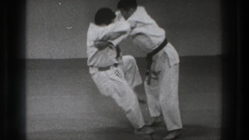 During the Tokyo practice sequence, one martial artist shifts balance and raises a knee while maintaining the grip.