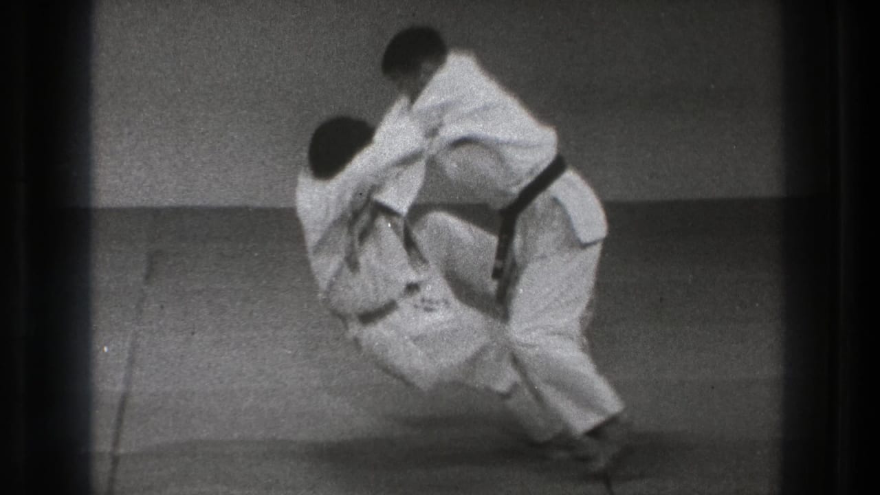 The 8mm frame shows the throw in progress, with one practitioner lowering toward the mat as the other controls the grip.