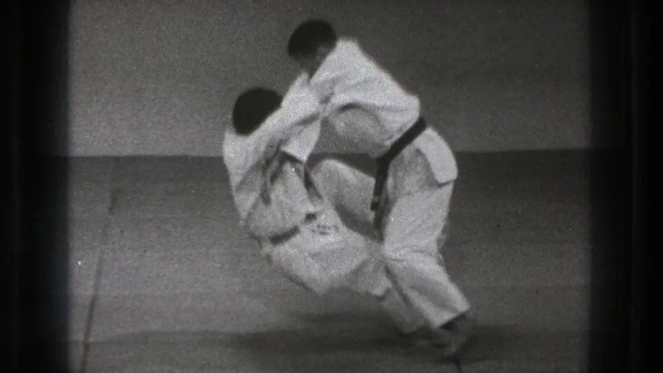 The 8mm frame shows the throw in progress, with one practitioner lowering toward the mat as the other controls the grip.