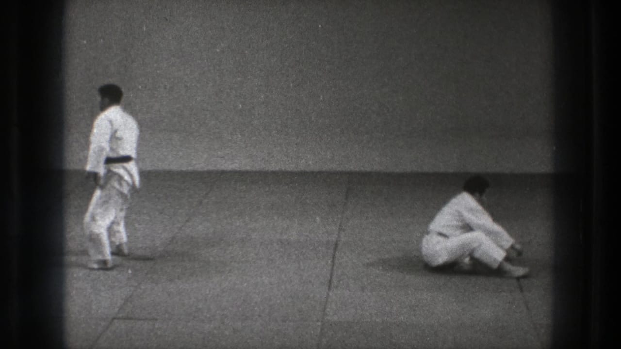 One man in a judogi stands on the dojo mat while another sits nearby in 1971 Tokyo judo practice footage.