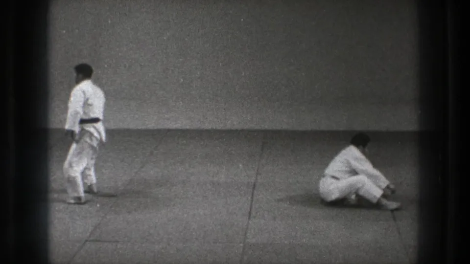 One man in a judogi stands on the dojo mat while another sits nearby in 1971 Tokyo judo practice footage.