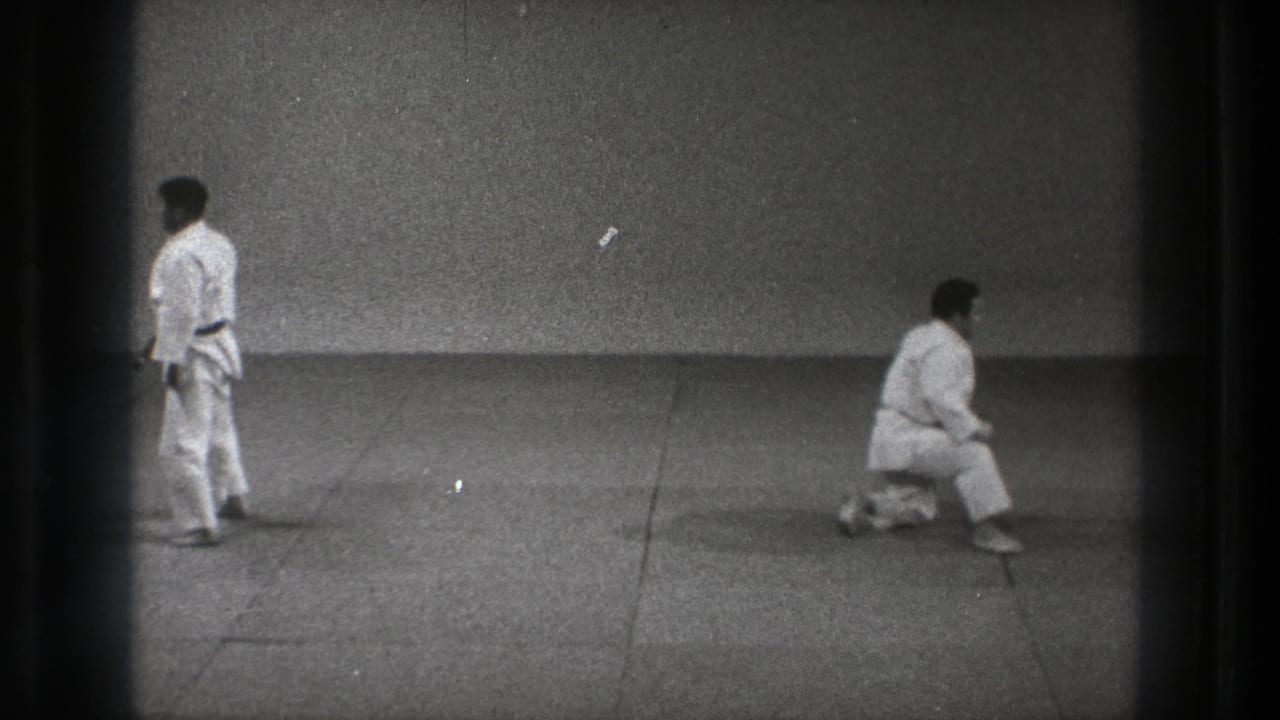 In Tokyo, 1971, two adult martial artists in white gi practice on a plain mat, one standing while the other crouches low.