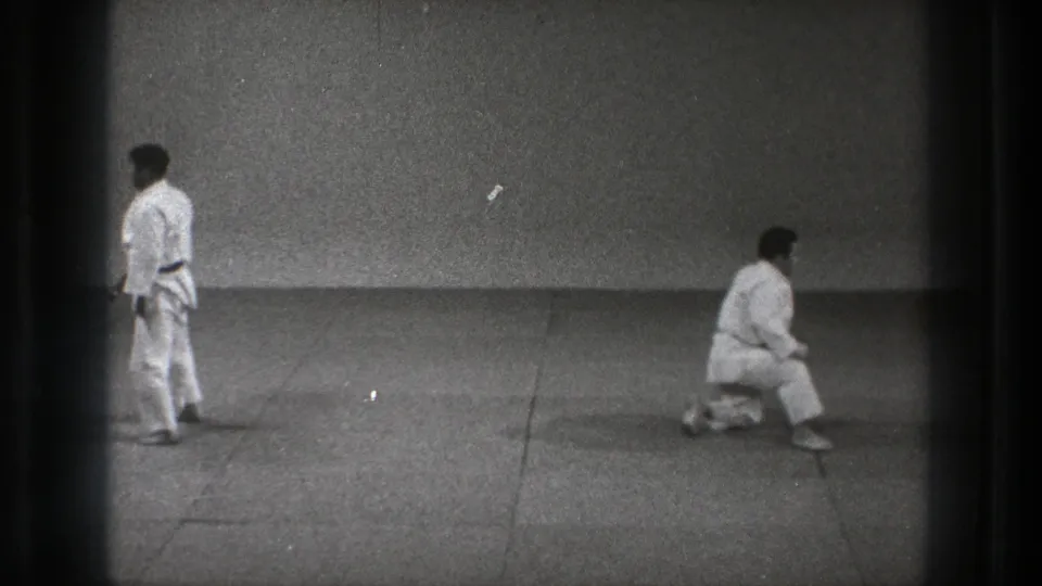 In Tokyo, 1971, two adult martial artists in white gi practice on a plain mat, one standing while the other crouches low.
