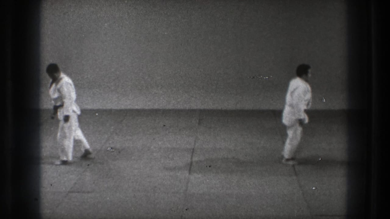 Two adult martial artists in white gi move apart inside a Tokyo dojo setting, filmed on grainy 8mm in 1971.