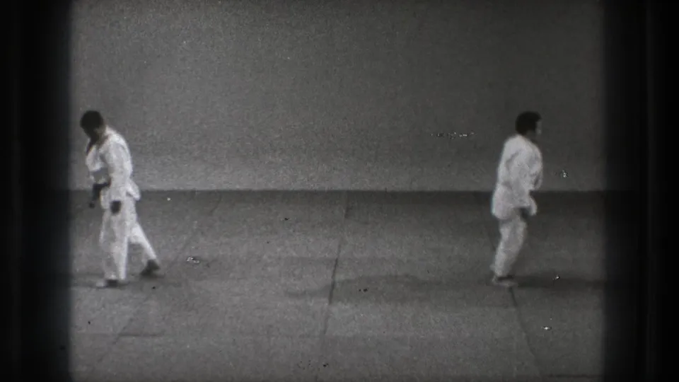 Two adult martial artists in white gi move apart inside a Tokyo dojo setting, filmed on grainy 8mm in 1971.