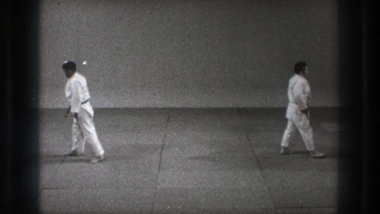 In a 1971 Tokyo dojo scene, two adults in white gi walk across the mat within a sparse training room.