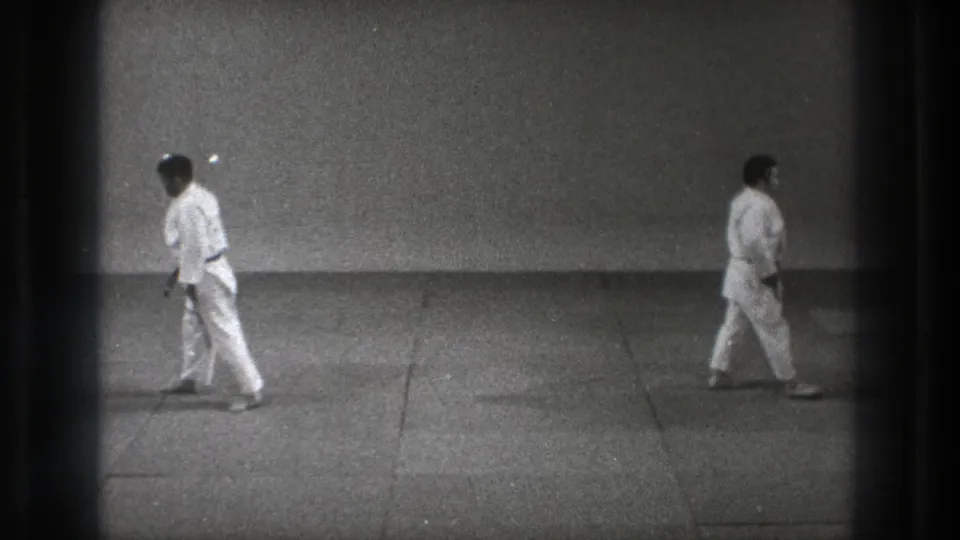 In a 1971 Tokyo dojo scene, two adults in white gi walk across the mat within a sparse training room.